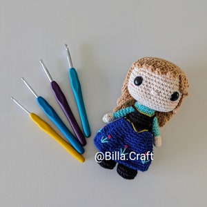 Op de afbeelding: Een gehaakte pop van een meisje met bruin haar, gekleed in een blauwe jurk met zwarte en gouden accenten. De pop ligt op een wit oppervlak met vier haaknaalden in verschillende kleuren.