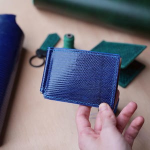 Puede incluir: Un monedero con clip para billetes de cuero azul con una superficie texturizada. El monedero se sostiene en una mano, con otras piezas de cuero y accesorios en el fondo. El monedero es de color azul oscuro.