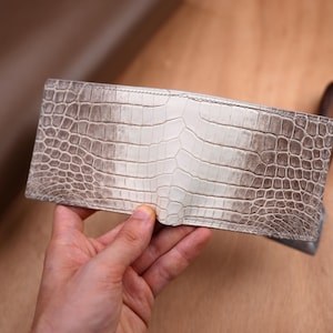 Op de afbeelding: Een bi-fold portemonnee met een gestructureerd, schubachtig patroon in een verloop van gebroken wit en lichtbruin. De portemonnee heeft een rechthoekige vorm met zichtbare stiksels langs de randen en een duidelijk herhalend reliëfpatroon.