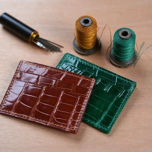 Könnte beinhalten: Zwei Leder-Kartenetuis, eines braun und eines grün, mit Krokodilleder-Muster, liegen auf einer Holzoberfläche. Zwei Garnrollen, gold und grün, sowie ein Nadelwerkzeug sind ebenfalls zu sehen.