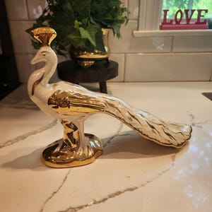May include: A white and gold ceramic peacock figurine. The peacock has a gold crown, gold accents on its wings and tail, and a gold base. The figurine is displayed on a white countertop.