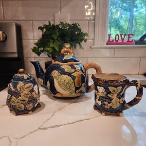 May include: A three-piece ceramic tea set with a navy blue background and a bird and pear design. The set includes a teapot, a sugar bowl, and a mug with a branch-like handle. The set is decorated with floral accents.