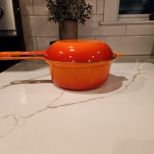 May include: A bright orange enameled cast iron saucepan with a matching lid. The pot features a long handle extending from the side and a smaller helper handle on the opposite side. The exterior has a smooth, glossy finish with a gradient color.
