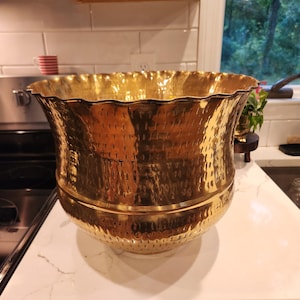 May include: A large, polished brass planter featuring a wide, scalloped rim and a textured, hammered finish across its exterior surface. The vessel has a rounded, bulbous body that tapers slightly toward the base, showcasing a reflective metallic gold tone.