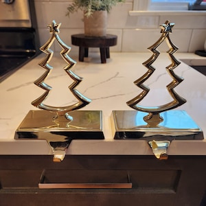 May include: Two gold-toned metal stocking holders, each featuring a cutout Christmas tree silhouette topped with a small star. The base of each holder is rectangular and includes a curved hook extending downward for hanging stockings.