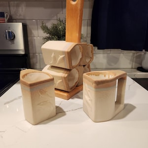 May include: A set of handmade ceramic mugs and a wooden holder. The mugs are off-white with a tan rim and a leaf design. The holder is made of light-colored wood. The mugs are arranged on a white countertop.