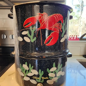 May include: A black enamel clam broth pot with a red lobster design and green seaweed accents. The pot has a spigot and the words "Clam Broth" are printed on the side. The pot is two-tiered and has handles.