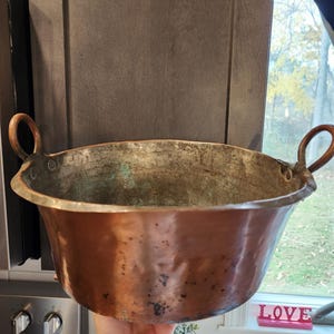 May include: A vintage copper bowl with two rounded handles. The bowl has a wide, shallow design with a slightly flared rim. The copper has a rich, aged patina with areas of green oxidation, showing its age and use.