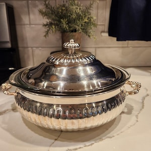 May include: A silver-plated serving dish with a lid. The dish has a round shape with a decorative, ribbed design around the base. The lid is topped with a decorative finial. The handles are ornate. The dish is on a white countertop.