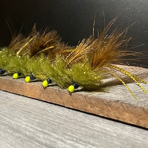 May include: A row of six green and brown fly fishing lures with yellow and black accents.