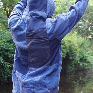 May include: A blue denim jacket with a patchwork design. The jacket has a hood and a dark blue pocket on the back. The jacket is worn with light blue denim shorts. The background is a blurred view of a river and trees.