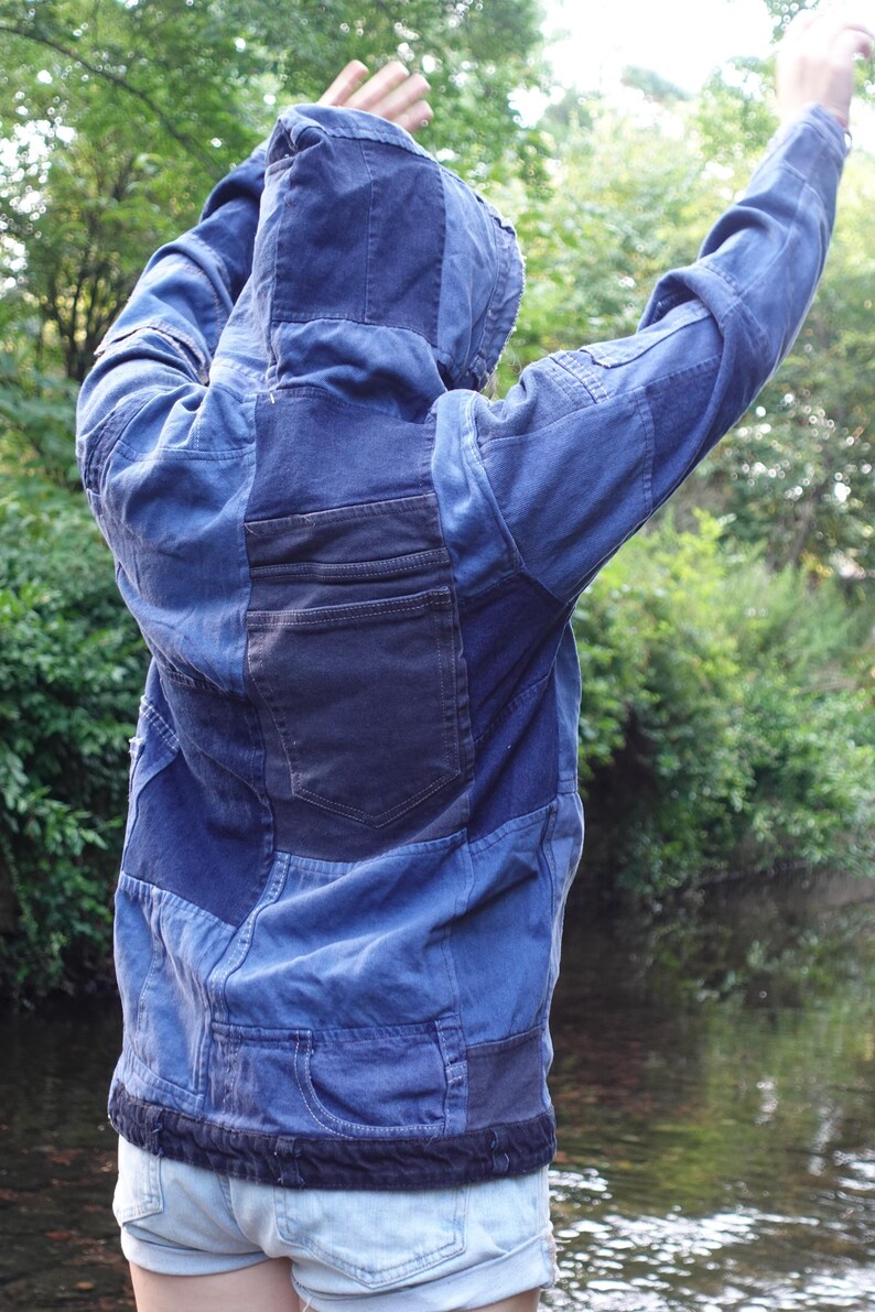 May include: A blue denim jacket with a patchwork design. The jacket has a hood and a dark blue pocket on the back. The jacket is worn with light blue denim shorts. The background is a blurred view of a river and trees.