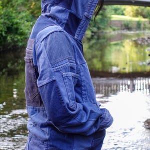 May include: A patchwork denim jacket in various shades of blue. The jacket features a hood and multiple panels sewn together, creating a unique design. The person is standing near a body of water.