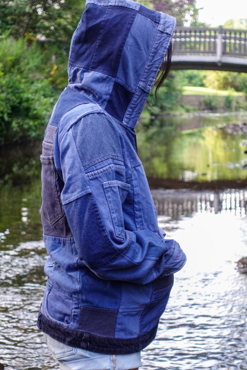 May include: A patchwork denim jacket in various shades of blue. The jacket features a hood and multiple panels sewn together, creating a unique design. The person is standing near a body of water.