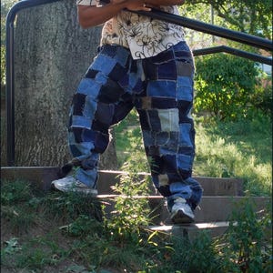 Puede incluir: Pantalones vaqueros azules de patchwork holgados. Los pantalones están hechos de cuadrados de mezclilla azul de varios tonos. La persona lleva zapatillas blancas. El fondo incluye un árbol, escaleras y vegetación.