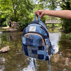 Puede incluir: Una mochila de mezclilla patchwork en varios tonos de azul. La mochila tiene un bolsillo frontal con cremallera beige y correas ajustables. La bolsa está sostenida por una mano frente a un río con un puente al fondo.