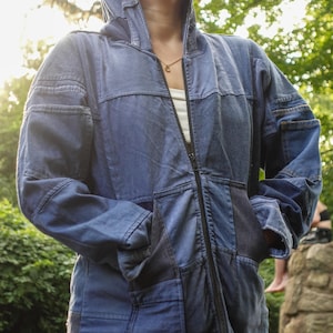 May include: A blue denim jacket with a patchwork design. The jacket has a hood, a full-length zipper, and pockets. The garment is worn over a white top.
