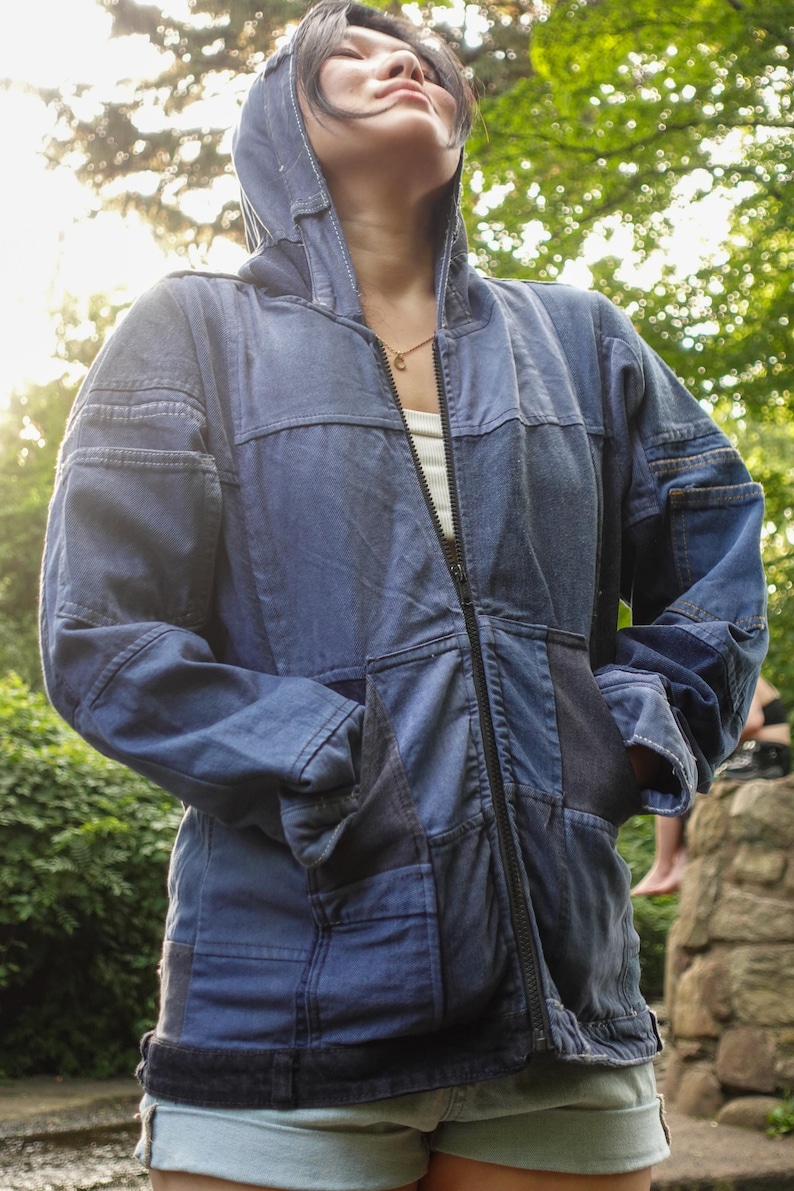 May include: A blue denim jacket with a patchwork design. The jacket has a hood, a full-length zipper, and pockets. The garment is worn over a white top.