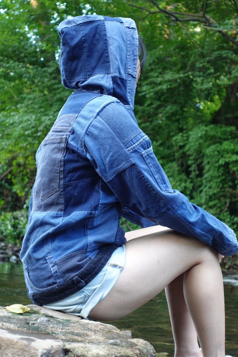 May include: A blue denim jacket with a patchwork design, featuring a hood and multiple shades of blue. The jacket has a pocket on the chest and is worn with light blue shorts. The person is sitting on a rock near a body of water.