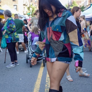 May include: A multi-colored patchwork corduroy jacket with a zipper closure. The jacket features a variety of colors including blue, red, pink, and brown. The person is wearing black boots and denim shorts. The background shows a street scene with other people.