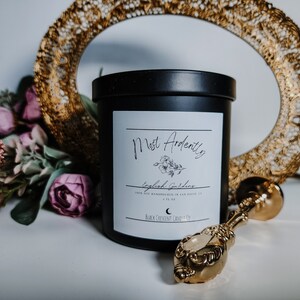 May include: Black candle jar with a white label that reads "Most Ardently English Gardens" with a small floral illustration. The candle is on a white surface with a gold decorative tray and a gold metal spoon.