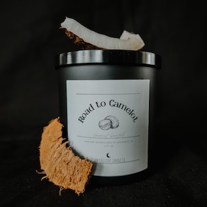 May include: Black candle jar with a white label that reads "Road to Camelot Coconut Sanfal 100% Soy Handpoured in San Diego, CA 8 fl oz Black Crescent Candle Co.". The label also features a drawing of two coconuts. The candle is sitting on a black surface with a piece of coconut shell next to it.