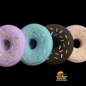 Op de afbeelding: Vier siliconen donutvormige kauwspeelgoed in paars, blauw, bruin en roze met witte en gekleurde sprinkels. De kauwspeelgoed zijn gerangschikt in een rij van links naar rechts.