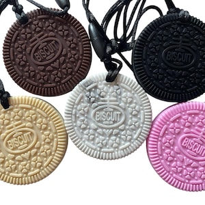 May include: Five circular pendants shaped like sandwich cookies, each hanging from a black cord. The pendants come in brown, black, beige, white, and pink. Each cookie features a textured floral pattern and the word BISCUIT embossed in the center.