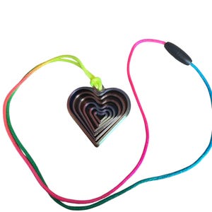 Puede incluir: Un collar con un colgante de corazón en capas y un cordón arcoíris. El corazón está hecho de múltiples formas de corazón anidadas en tonos marrones y grises. El cordón pasa de verde a rosa a azul, con un cierre negro.