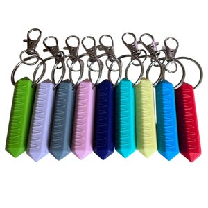 Op de afbeelding: Een verzameling kleurrijke sleutelhangers met geometrische, prismavormige hangers. De sleutelhangers hebben een verscheidenheid aan kleuren, waaronder groen, lavendel, grijs, roze, marineblauw, turquoise, geel, blauw en rood. Elke sleutelhanger heeft een zilverkleurige sluiting en ring.