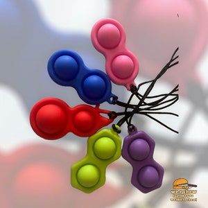 May include: Five colorful pop it fidget toys with black strings attached. The toys are in the following colors: pink, blue, red, green, and purple.