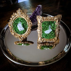 Puede incluir: Dos marcos dorados ornamentados, uno ovalado y otro rectangular, cada uno con musgo verde y un cristal de cuarzo transparente. Los marcos están sobre una bandeja plateada, con un cristal de amatista púrpura detrás. Estética vintage.