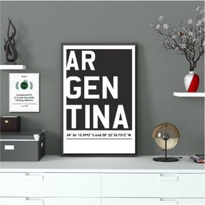 Könnte beinhalten: Gerahmter Schwarz-Weiß-Druck mit dem Wort "ARGENTINA" in großen weißen Großbuchstaben. Darunter befindet sich eine Textzeile mit den Koordinaten 34° 36' 13.3992" S und 58° 22' 53.7312" W. Der Druck befindet sich auf einer weißen Oberfläche.