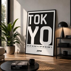 Könnte beinhalten: Ein vertikales, rechteckiges Wandbild mit dem Wort TOKYO in großen, weißen serifenlosen Buchstaben auf schwarzem Hintergrund. Unter dem Text sind die Koordinaten 35° 39' 10.1952" N, 139° 50' 22.1208" E und das Wort JAPAN aufgedruckt.