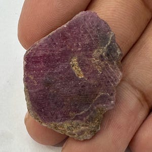 Puede incluir: Una piedra preciosa de rubí en bruto y sin pulir con un tono magenta y púrpura intenso. La piedra presenta una forma irregular y dentada con una superficie texturizada y mate, además de inclusiones cristalinas visibles en su estructura.