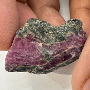 Puede incluir: Un espécimen mineral en bruto y sin pulir que presenta una estructura cristalina de color magenta rosado vibrante incrustada en una matriz de color gris verdoso oscuro y texturizado. La piedra tiene una forma irregular y dentada con estrías visibles y una superficie natural rugosa.
