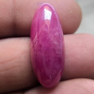 Può includere: Una gemma lucidata, di forma ovale, con una vibrante tonalità rosa. La pietra ha una superficie liscia e lucida e mostra motivi interni di rosa chiaro e bianco. La gemma è tenuta tra le dita, mostrando le sue dimensioni e il suo colore.