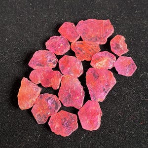 May include: A cluster of rough, pink gemstones. The stones are of varying sizes and shapes, with a translucent appearance. The colour varies from a bright pink to a deeper reddish-pink. The gemstones are presented on a dark, textured surface.