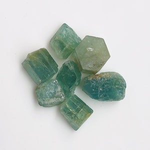 Puede incluir: Un grupo de seis cristales de aguamarina, cada uno con una forma y un tamaño únicos. Los cristales son de color azul verdoso claro y tienen una apariencia translúcida.