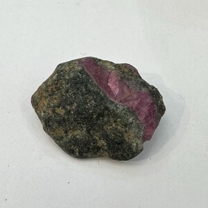 Op de afbeelding: Een ruw, ongepolijst mineraal exemplaar met een donkere, getextureerde, grijszwarte rotsmatrix met een opvallend, ingebed kristallijn gedeelte van levendig, doorschijnend magenta-roze korund, met natuurlijke grillige randen en oppervlakken.