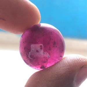 May include: A round, polished ruby gemstone held between fingers. The gem is a deep, translucent pink with darker inclusions, reflecting light. The background is a bright blue sky.