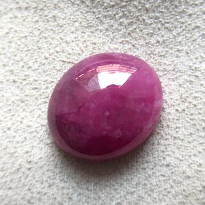 Könnte beinhalten: Ein oval geschliffener Rubin-Edelstein. Der Edelstein hat eine tiefe, gesättigte, rötlich-rosa Farbe mit einer glänzenden, reflektierenden Oberfläche. Der Stein liegt auf einem strukturierten, cremefarbenen Hintergrund.