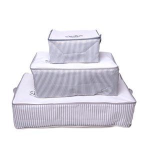 Puede incluir: Tres cajas de almacenamiento blancas apiladas, cada una con un patrón de rayas grises y blancas. Las cajas tienen ribetes y cremalleras grises. La parte superior de cada caja tiene un pequeño diseño bordado.