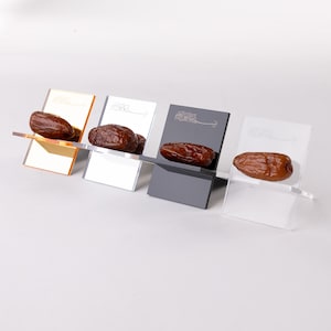 May include: Four small, angled display stands in gold, silver, gray, and clear acrylic, each holding a single brown date. The stands have a minimalist design, with a slot to hold the date. The background is white.