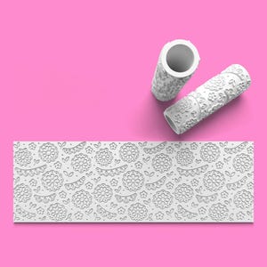 May include: Two white cylindrical paint rollers featuring an embossed floral pattern and a flat rectangular strip displaying the same repeating design of flowers, leaves, and scalloped shapes in a white relief on a white background.