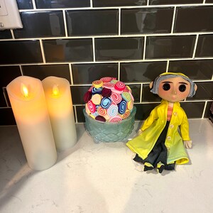 May include: A colourful fabric ball sits in a blue glass bowl, next to two white candles and a doll wearing a yellow raincoat. The doll has grey hair and large eyes.