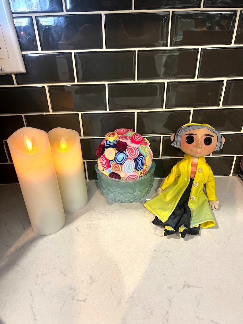 May include: A colourful fabric ball sits in a blue glass bowl, next to two white candles and a doll wearing a yellow raincoat. The doll has grey hair and large eyes.