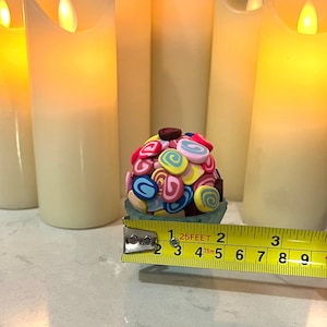 May include: A colorful, multi-colored, swirl-shaped candy ball in a clear plastic container, measured with a yellow tape measure that reads "25 FEET".