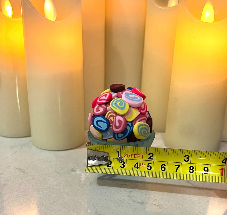 May include: A colorful, multi-colored, swirl-shaped candy ball in a clear plastic container, measured with a yellow tape measure that reads "25 FEET".