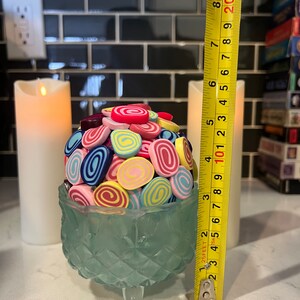 May include: A teal glass pedestal with three legs holds a cluster of colourful, swirl-shaped sweets. The sweets are in shades of pink, blue, yellow, and green. A yellow measuring tape is visible to the right of the pedestal.